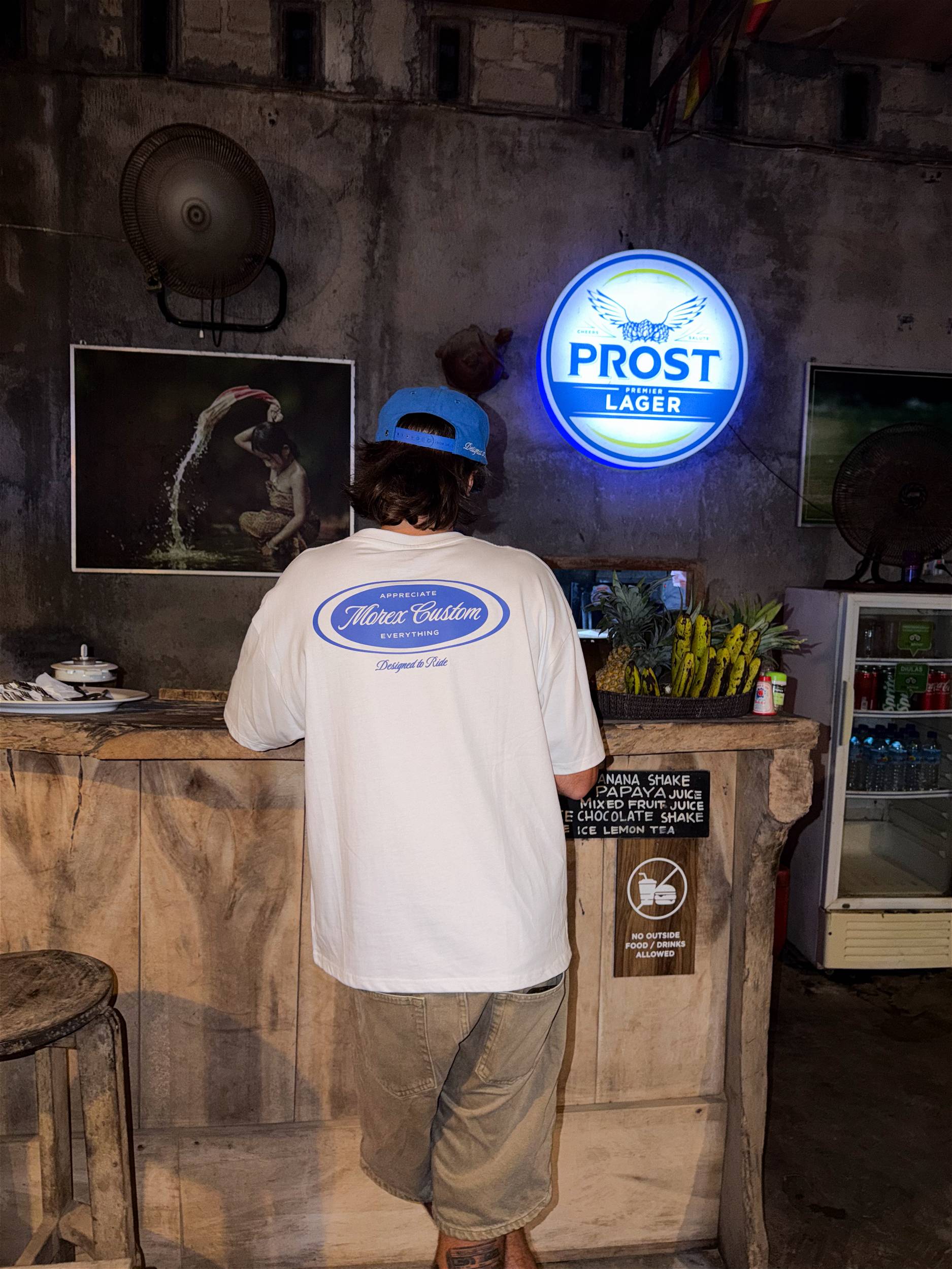 Un homme de dos portant un t-shirt blanc Morex Custom avec un logo bleu imprimé au dos, se tenant devant un bar au style industriel avec une enseigne néon Prost Beer.