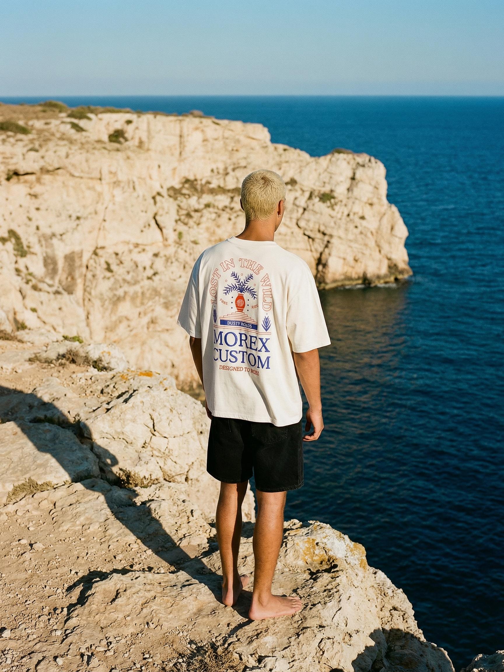 Homme de dos portant un t-shirt blanc Morex Custom avec un design graphique, debout sur une falaise rocheuse face à l'océan bleu.