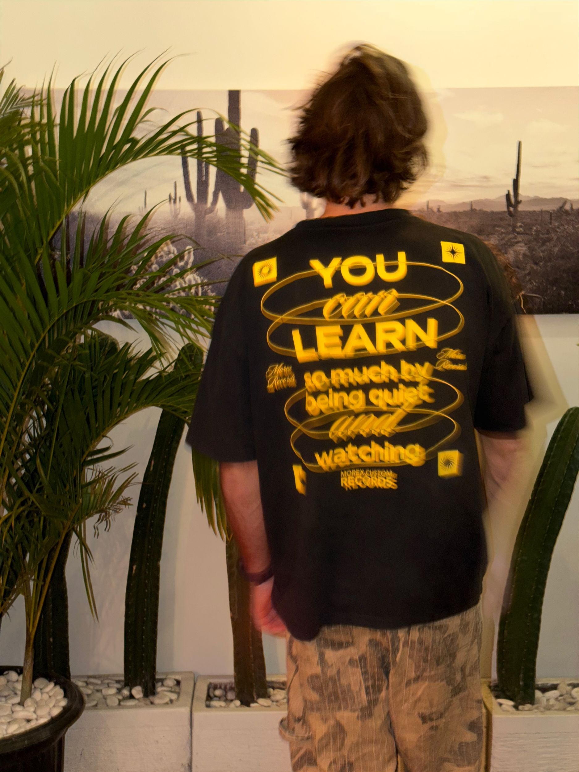 T-shirt noir Morex Custom avec typographie jaune 'You can learn so much by being quiet and watching', porté par un homme dans un décor lifestyle avec cactus.
