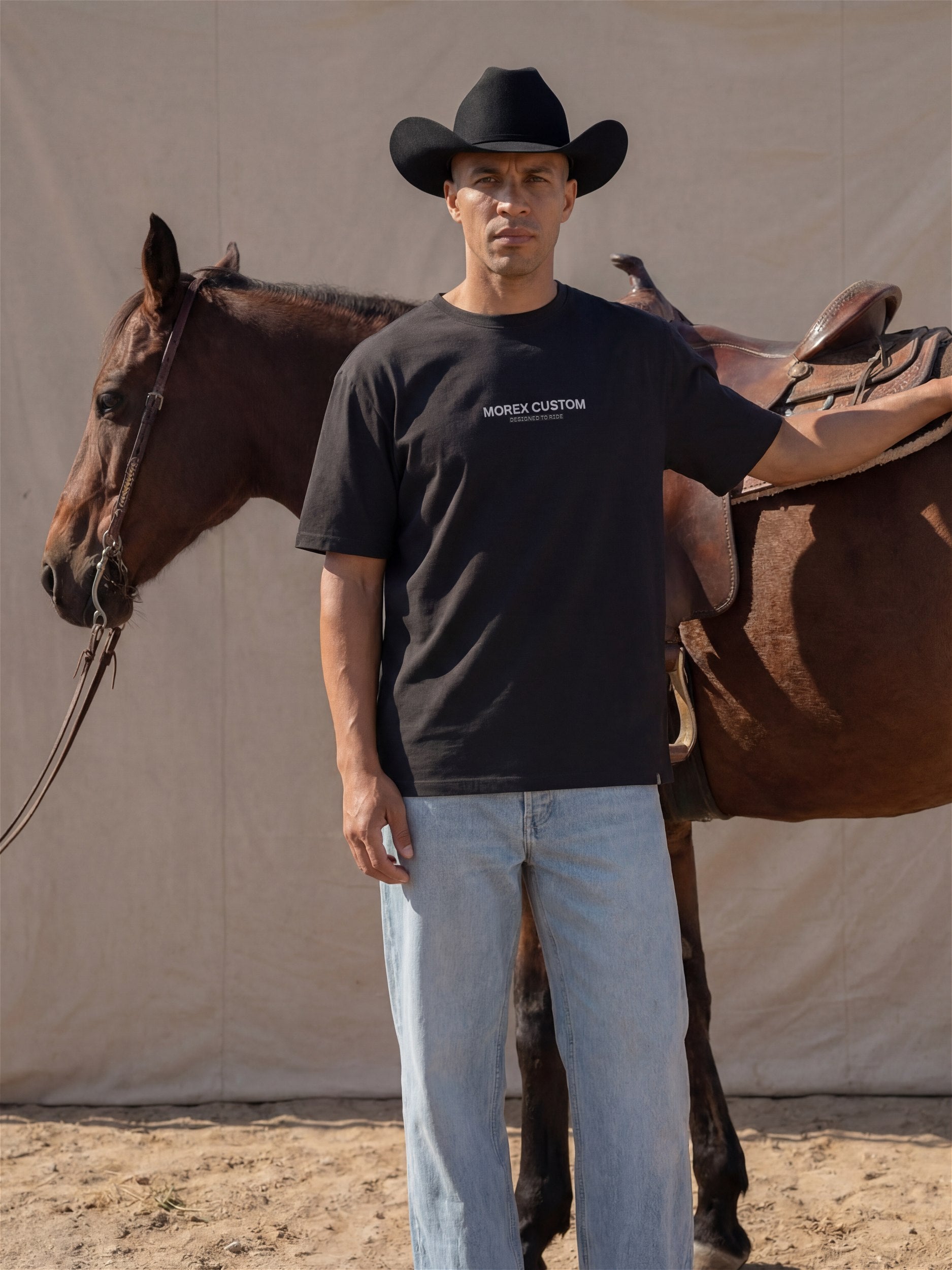 Homme portant un t-shirt Morex Custom noir et un chapeau de cowboy, posant à côté d'un cheval brun sellé dans un style lifestyle western.