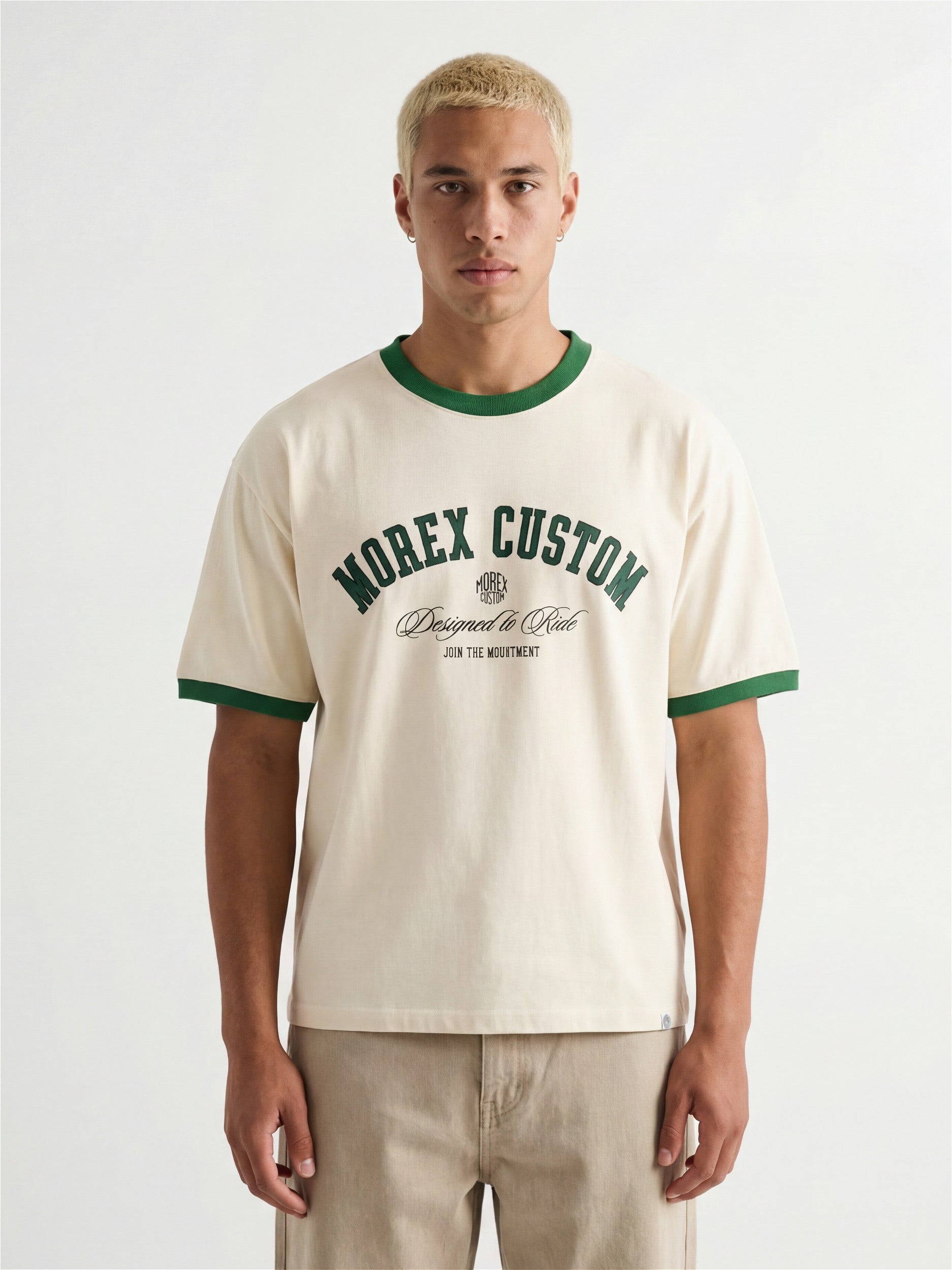 Un homme portant un t-shirt ringer Morex Custom de couleur crème avec des bordures vertes au col et aux manches, affichant un logo graphique de style moto lifestyle.